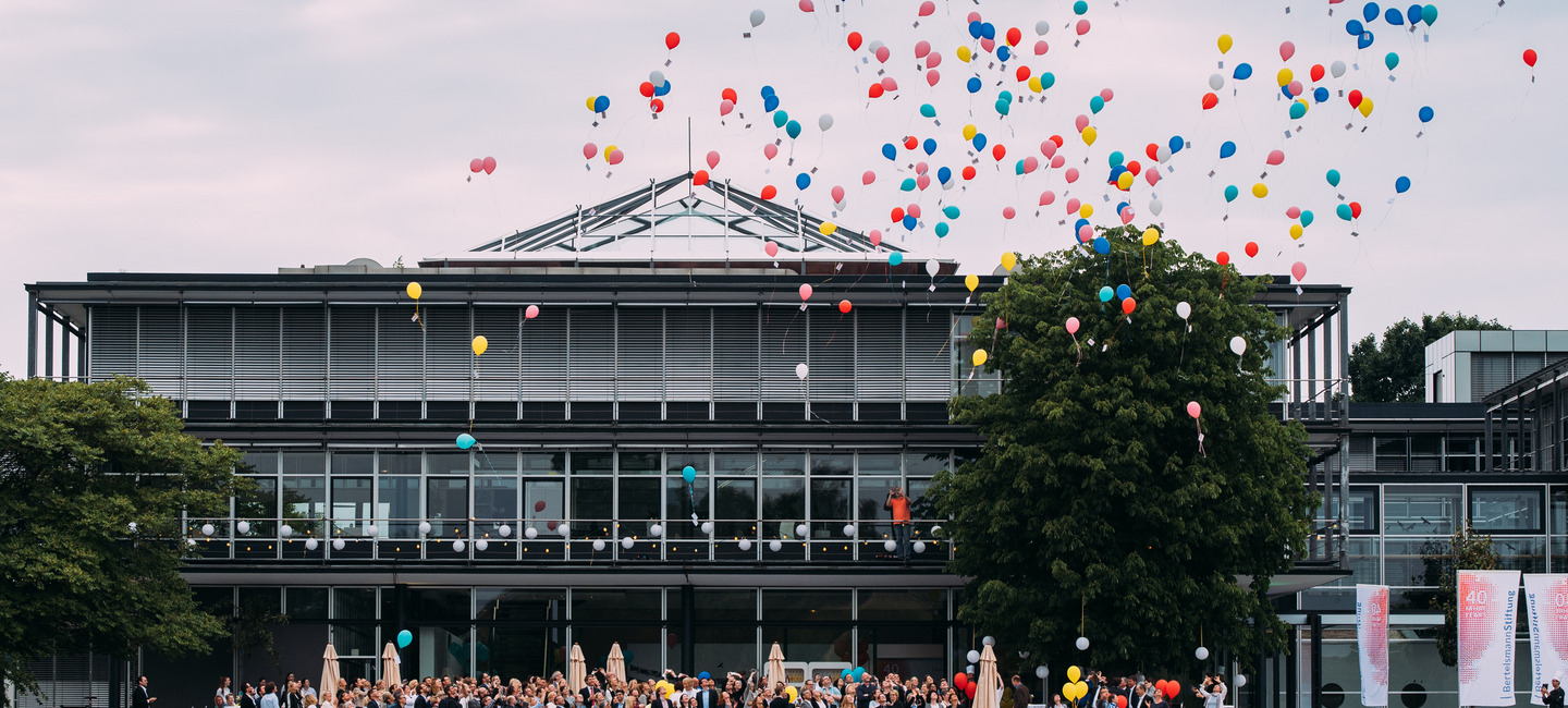 Blick über den See vor dem Gebäude auf das Gebäude der Bertelsmann Stiftung während eines Sommerfestes. Bunte Luftballons steigen von der Terrasse vor dem Gebäude auf, auf der die Mitarbeiter:innen der Stiftung versammelt sind.
