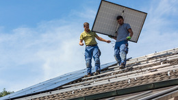 Zwei Männer laufen über ein Dach, vom First Richtung Dachrinne. Einer von beiden trägt ein Solarpanel.