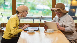 [Translate to English:] Zwei ältere Frauen sitzen am Tisch. Die eine Frau sitzt vor einem Laptop und schaut gespannt auf den Bildschirm. Die zweite Frau hat ein Tablet in der Hand und schaut auf dieses. Beide Frauen sehen glücklich aus uns schmunzeln.