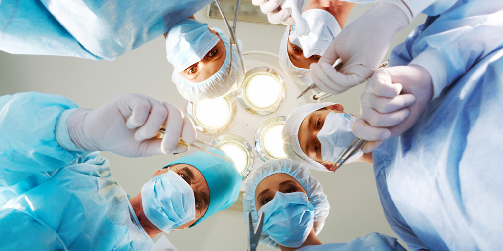 Aus der Perspektive eines im Operationssaal liegenden Patienten sind fünf Ärzte in ihren Operationskitteln und mit Mundschutz zu sehen, die sich über den Patienten beugen und Scheren und Skalpelle in der Hand haben. Sie blicken dem Patienten in die Augen.