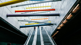 [Translate to English:] Rolltreppen in einer U-Bahn-Station. Der Blick richtet sich an die Decke wo in bunten Farben Verbindunsbalken zwischen der rechten und linken Wand zu sehen sind.