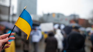 eine Hand mit einer Ukrainischen Flagge im Hintergrund stehen Menschen