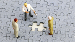 drei Modellfiguren stehen auf einer grauen Puzzlefläche, ein Puzzleteil fehlt in der Mitte