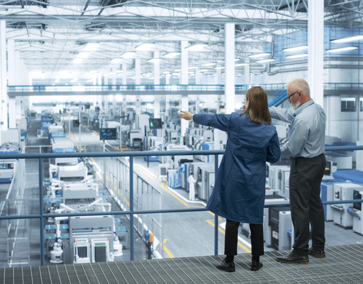 Eine Ingenieurin und ein Ingenieur stehen mit dem Rücken zur Kamera auf einem Podest und besprechen die Produktion in einer modernen, automatisierten Elektronikfertigung.