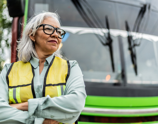 Eine grauhaarige Frau steht als Busfahrerin mit Warnweste bekleidet vor einem Bus.