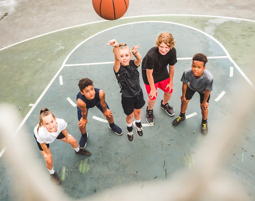 Fünf Kinder stehen auf einem Basketballfeld vor dem Korb. Eines der Kinder wirft den Ball in Richtung Korb.