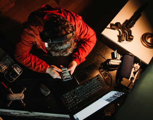 Eine Person mit Kopfhörern und roter Jacke sitzt an einem Schreibtisch und blickt auf ihr Smartphone. Um sie herum befinden sich ein Laptop, eine Kamera, ein Mikrofon und weitere technische Geräte. Die Aufnahme zeigt die Szene aus der Vogelperspektive bei gedämpftem Licht.