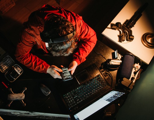 Eine Person mit Kopfhörern und roter Jacke sitzt an einem Schreibtisch und blickt auf ihr Smartphone. Um sie herum befinden sich ein Laptop, eine Kamera, ein Mikrofon und weitere technische Geräte. Die Aufnahme zeigt die Szene aus der Vogelperspektive bei gedämpftem Licht.