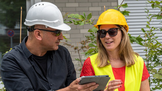 Zwei Arbeiter auf einer Baustellen tragen Sicherheitskleidung und -weste und prüfen gemeinsam etwas auf einem Tablet.