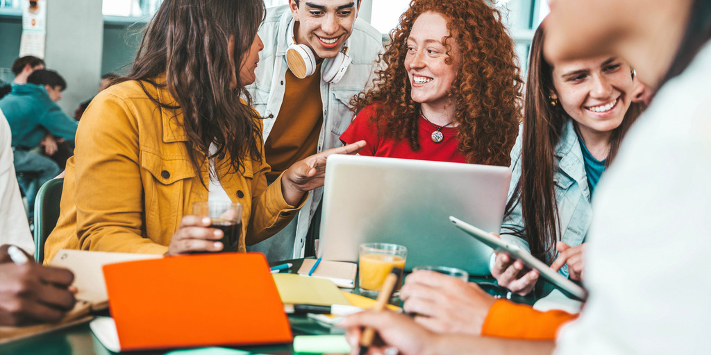 Fünf junge Menschen sitzen arbeitend an einem Tisch beisammen. Es liegen Laptops und Schreibblöcke vor ihnen.