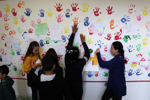 Kinder machen bunte Abdrücke ihrer Hände an eine weiße Wand und schreiben etwas dazu auf.