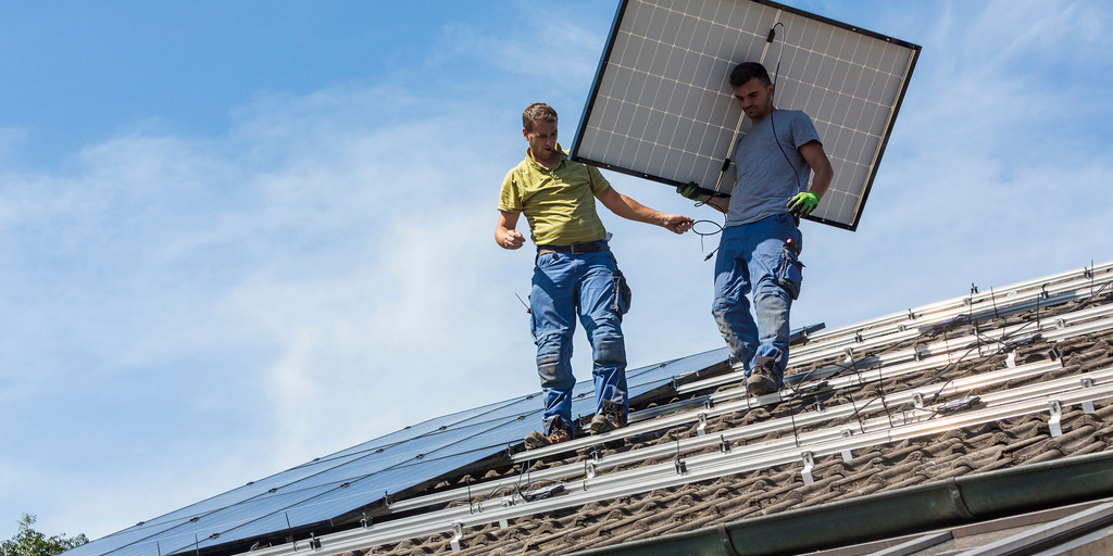 Zwei Männer laufen über ein Dach, vom First Richtung Dachrinne. Einer von beiden trägt ein Solarpanel.