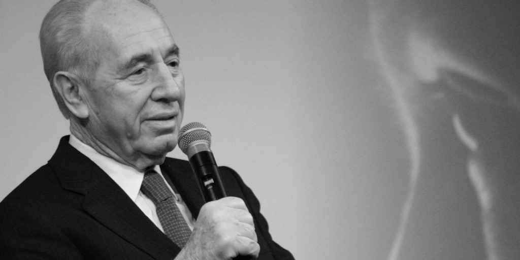 Shimon Peres, ehemaliger Premier und Staatspräsident Israels, sitzt während einer Veranstaltung auf einem Podium und hält ein Mikro in der Hand. Im Hintergrund ist eine Leinwand mit dem Ausschnitt eines Gesichts zu sehen.