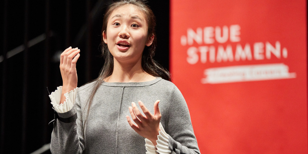 The young opera singer Chen Wang from China sings in the NEUE STIMMEN Master Class. The black-haired young woman stands in front of a red NEUE STIMMEN banner.