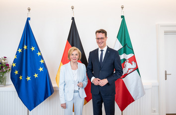 Liz Mohn und Hendrik Wüst stehen für ein gemeinsames Foto in der Staatskanzlei des Landes Nordrhein-Westfalen vor Fahnen der Europäischen Union, Deutschlands und des Landes Nordrhein-Westfalen.