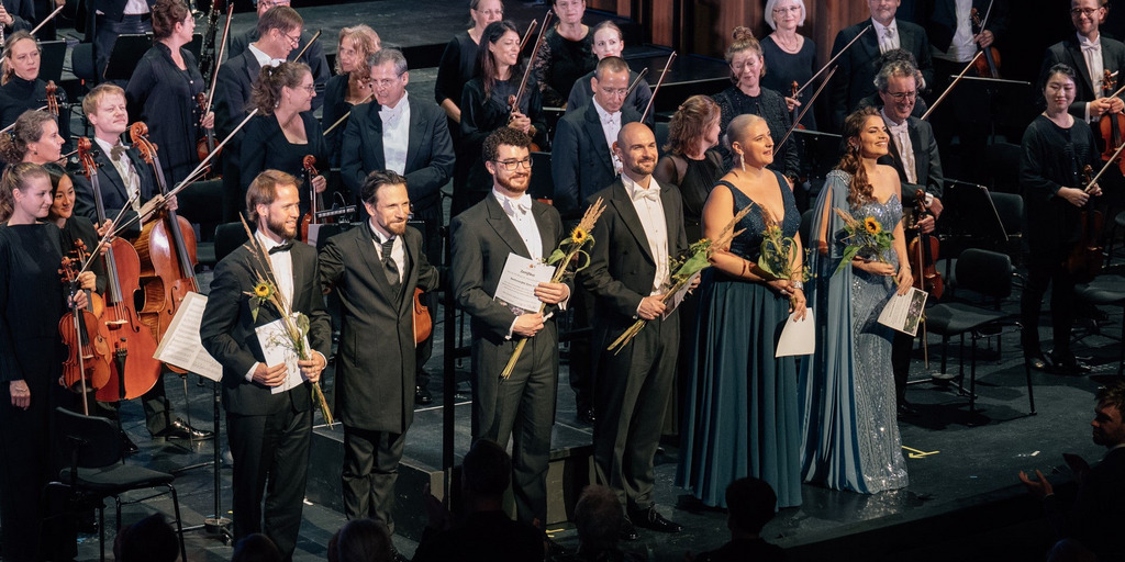 Die Solist:innen, das Orchester, der Dirigent und der Leiter des Chores stehen auf der Bühne der Oper Bonn und nehmen den Applaus des Publikums entgegen.