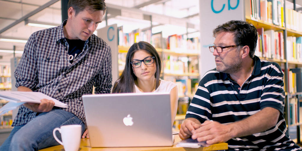 [Translate to English:] Drei Personen in einer Arbeitsgruppe vor einem Laptop in einer Bibliothek.