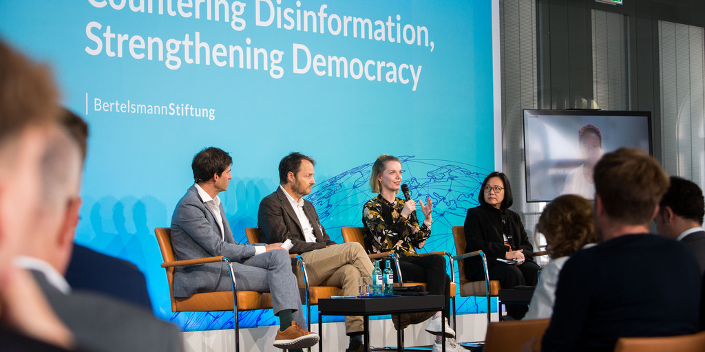 A look onto the stage during the panel discussion "Democracy in Times of Disinformation". Cathleen Berger from Bertelsmann Stiftung is sitting on a chair and speaking. Next to her, moderator Dominik Hierlemann from Bertelsmann Stiftung, Michael Meyer-Resende from Democracy Reporting International and Yvonne Chua from the University of the Philippines are sitting on chairs and listening to Cathleen Berger.