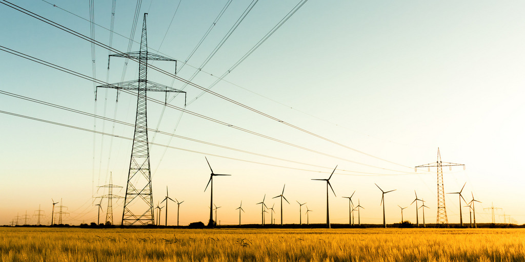 The picture shows a landscape with several high-voltage pylons carrying power lines. There are numerous wind turbines in the background. The power lines run across the picture, while the wind turbines are located in a flat, open environment.