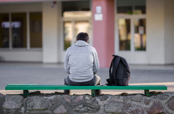 Ein junger Mann sitzt alleine auf einer Bank. Neben ihm steht ein Rucksack. Er sitzt gebückt.
