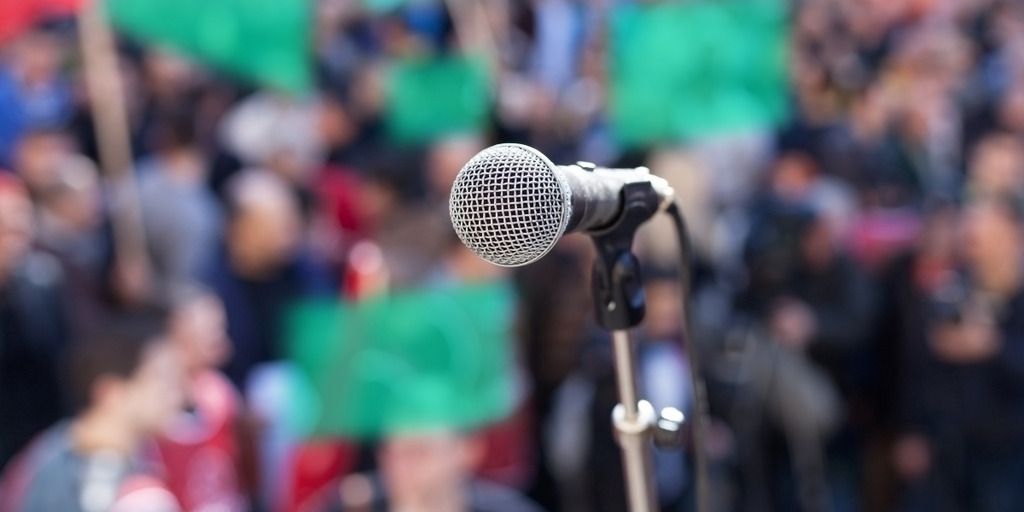 Vor einer Menschenmenge, die grüne Plakate in die Höhe hält, ist ein Mikrofon auf einer Bühne zu sehen, vor dem noch kein Redner steht.