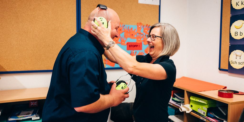 Christiane Godt, Vorsitzende der Gütersloher Stadtschulpflegschaft, setzt ihrem Ko-Vorsitzenden Christian Beckmann in einem Schulraum Kopfhörer zum Schutz seiner Ohren auf und lacht.