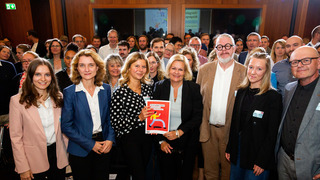 Gruppenfoto von Bundesinnenministerin Nancy Faeser, Daniela Schwarzer, Vorständin der Bertelsmann Stiftung, und Teilnehmenden des Bürgerrates bei der Übergabe des Bürgergutachtens zum "Forum gegen Fakes". Nancy Faeser und eine Teilnehmende des Bürgerrates werden von den anderen Teilnehmenden umgeben und halten lächelnd das Bürgergutachten in die Kamera.