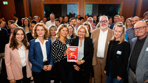 Gruppenfoto von Bundesinnenministerin Nancy Faeser, Daniela Schwarzer, Vorständin der Bertelsmann Stiftung, und Teilnehmenden des Bürgerrates bei der Übergabe des Bürgergutachtens zum "Forum gegen Fakes". Nancy Faeser und eine Teilnehmende des Bürgerrates werden von den anderen Teilnehmenden umgeben und halten lächelnd das Bürgergutachten in die Kamera.