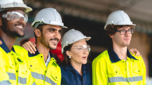 Drei Männer und eine Frau in Arbeitskleidung, mit Bauhelmen auf dem Kopf, blicken in die Ferne und lächeln.