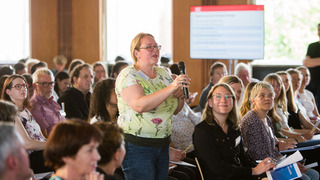 Eine Teilnehmerin steht bei einem Treffen des Bürgerrates für das Forum gegen Fakes im Saal und spricht in ein Mikrofon, während ihr andere Teilnehmer:innen zuhören.