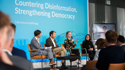 A look onto the stage during the panel discussion "Democracy in Times of Disinformation". Cathleen Berger from Bertelsmann Stiftung is sitting on a chair and speaking. Next to her, moderator Dominik Hierlemann from Bertelsmann Stiftung, Michael Meyer-Resende from Democracy Reporting International and Yvonne Chua from the University of the Philippines are sitting on chairs and listening to Cathleen Berger.