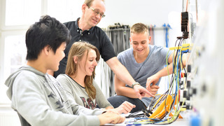 Das Bild zeigt eine Szene in einem Klassenzimmer oder Schulungsraum, in dem vier Personen interagieren. Im Vordergrund sitzen drei junge Erwachsene, zwei Männer und eine Frau, an einem Tisch mit verschiedenen elektrischen Messgeräten und Kabeln. Alle drei lächeln und sind offensichtlich in ein gemeinsames Projekt vertieft. Hinter ihnen steht eine ältere Person, vermutlich der Lehrer oder Ausbilder, der auf eines der Geräte zeigt und den Jugendlichen etwas erklärt. Die Atmosphäre wirkt konzentriert und gleichzeitig freundlich.