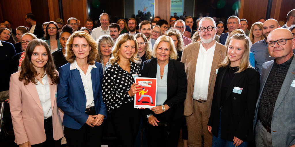 Gruppenfoto von Bundesinnenministerin Nancy Faeser, Daniela Schwarzer, Vorständin der Bertelsmann Stiftung, und Teilnehmenden des Bürgerrates bei der Übergabe des Bürgergutachtens zum "Forum gegen Fakes". Nancy Faeser und eine Teilnehmende des Bürgerrates werden von den anderen Teilnehmenden umgeben und halten lächelnd das Bürgergutachten in die Kamera.