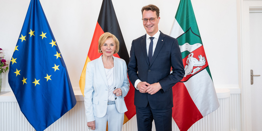 Liz Mohn und Hendrik Wüst stehen für ein gemeinsames Foto in der Staatskanzlei des Landes Nordrhein-Westfalen vor Fahnen der Europäischen Union, Deutschlands und des Landes Nordrhein-Westfalen.