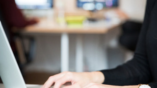 Eine Person in einem Büro tippt auf einer Laptop-Tastatur. Im Hintergrund sitzen weitere Personen an Bildschirmen.