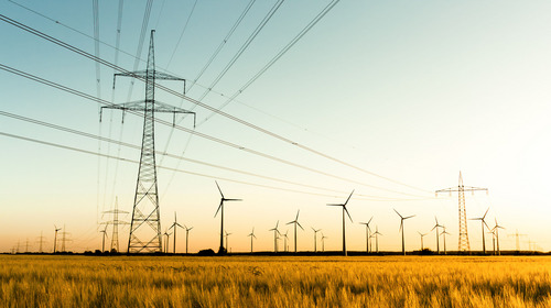 Das Bild zeigt eine Landschaft mit mehreren Hochspannungsmasten, die Stromleitungen tragen. Im Hintergrund stehen zahlreiche Windkraftanlagen. Die Stromleitungen verlaufen quer über das Bild, während sich die Windräder in einer flachen, offenen Umgebung befinden.