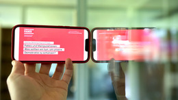 Ein Handy wird schräg gegen ein Spiegel gehalten. Auf dem Display sieht man die Schrift "Fakes und Manipulation: Was sollten wir tun, um unsere  Demokratie zu schützen?"