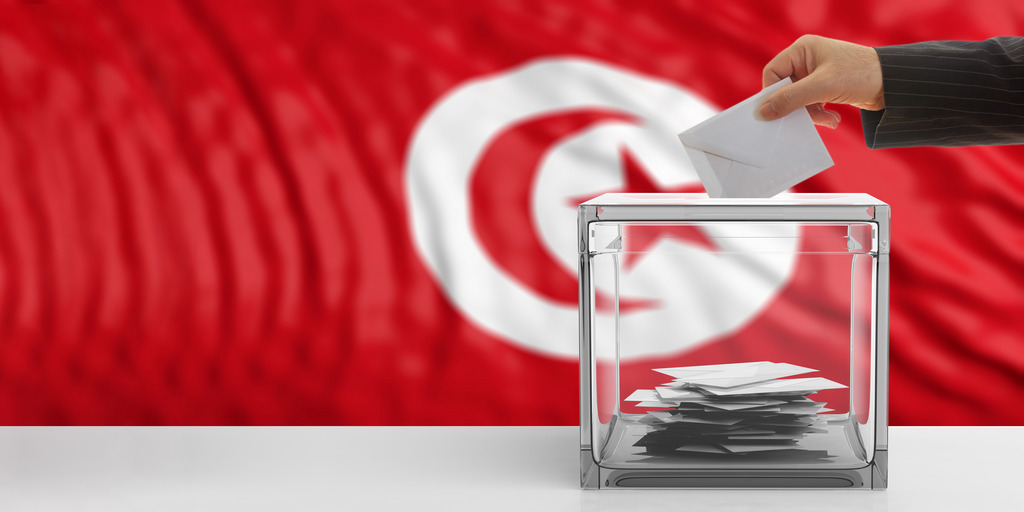 Hand wirft einen Stimmzettel in Wahlurne vor tunesischer Fahne
